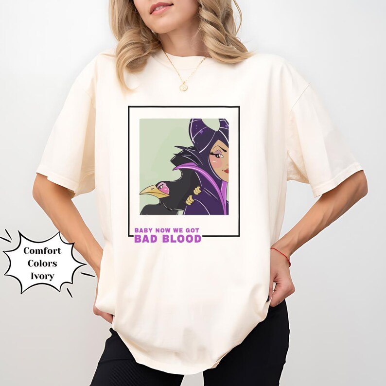 May include: A white t-shirt with a graphic print of Maleficent from Disney's Sleeping Beauty. The graphic is in a black and purple color scheme and features Maleficent with her raven. The text "Baby Now We Got Bad Blood" is printed below the graphic.