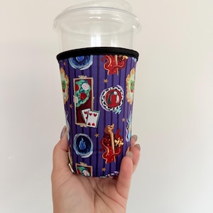 May include: A clear plastic cup with a fabric sleeve featuring a purple background with vertical black stripes. The sleeve is decorated with various colourful icons including playing cards, mirrors, flames, and circular emblems with detailed designs.