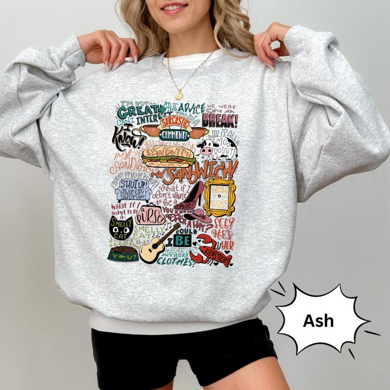 Op de afbeelding: Een grijs sweatshirt met een witte print van verschillende zinnen en afbeeldingen uit de populaire tv-serie "Friends". De print bevat de woorden "Het spijt me", "we waren uit elkaar", "hoe gaat het?" en "mijn sandwich".
