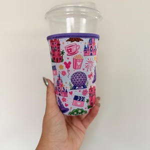 May include: A clear plastic cup with a fabric sleeve featuring a repeating pattern of pink and purple theme park icons, including castles, a geodesic dome, teacups, popcorn, fireworks, and Mickey Mouse silhouettes on a light blue background.