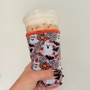 May include: A pink and orange Halloween-themed drink sleeve with a floral and spooky pattern. The design includes ghosts, bats, and pumpkins. The sleeve is wrapped around a clear plastic cup filled with a beverage.
