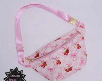May include: Pink fanny pack with a cherry and bow pattern on a white and pink checkered background. The bag has a pink adjustable strap and gold-colored hardware. The text "THAT'S TOO GRAPHIC, LLC" is in a black starburst.