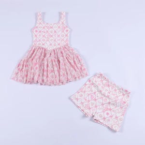 Può includere: Un completo da bambino composto da un vestito senza maniche e pantaloncini abbinati. Entrambi gli articoli presentano un motivo rosa e bianco. L'abito ha una gonna a balze. L'outfit è adatto per le giornate calde.