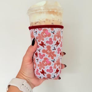 May include: A light purple drink sleeve with a festive gingerbread man and castle pattern. The sleeve is wrapped around a clear plastic cup filled with an iced beverage. Red trim accents the top edge of the sleeve.