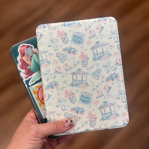 以下が含まれることがあります： スケート、リボン、ガゼボを特徴とする冬をテーマにしたパターンが施された、明るい色の電子書籍リーダーケース。ケースは手に持っており、人魚のステッカーがターコイズ色のケースに貼られているのが見えます。