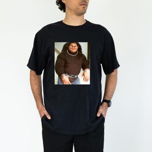 Op de afbeelding: Een zwart T-shirt met een bedrukte afbeelding van een man met een groot hoofd en een gouden ketting. De man draagt een bruine turtleneck, een blauwe jeans en een zwarte heuptas.