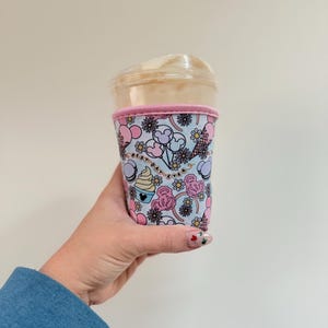 以下が含まれることがあります： パステルカラーの風船、花、カップケーキの模様が施された、再利用可能なコーヒーカップスリーブ。スリーブはピンクのトリムで、「Best Day Ever」の文字が見えます。透明なプラスチックカップに巻かれています。