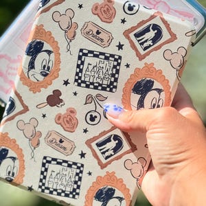 Op de afbeelding: Crèmekleurige tablethoes met een herhalend patroon van Mickey Mouse-portretten, ballonnen en omlijnde silhouetten. Het ontwerp bevat het woord "Dream" op een ticketvormige afbeelding, samen met sterren en een kasteelillustratie. De hoes wordt in een hand gehouden.