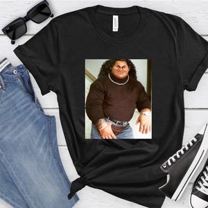 Op de afbeelding: Zwart T-shirt met een afbeelding van een man die een zwarte turtleneck en een blauwe jeans draagt. De man heeft een dikke snor en baard.