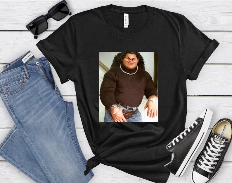 Op de afbeelding: Zwart T-shirt met een afbeelding van een man die een zwarte turtleneck en een blauwe jeans draagt. De man heeft een dikke snor en baard.