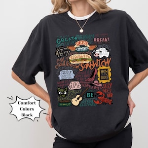 Op de afbeelding: Zwart T-shirt met een kleurrijke grafische print van verschillende zinnen en afbeeldingen uit de populaire tv-serie "Friends". De grafiek bevat de tekst "Ik ben niet goed in dit", "Hoe gaat het?", "Draai", "Stinkende kat", "Mijn sandwich" en "We waren in een pauze!"