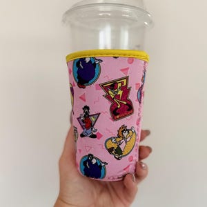 May include: A pink neoprene cup sleeve featuring a repeating pattern of cartoon characters and the text Powerline, accented with a yellow top trim, wrapped around a clear plastic cup with a matching clear plastic lid for cold beverages.