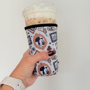 Könnte beinhalten: Ein weiß-schwarzer Getränkebecher mit Mickey Mouse in einem goldenen Rahmen. Der Becher hat eine schwarze Einfassung und ist um einen transparenten Plastikbecher gewickelt, der mit einem Getränk und Schlagsahne gefüllt ist. Das Wort "Dream" ist auf dem Becher aufgedruckt.