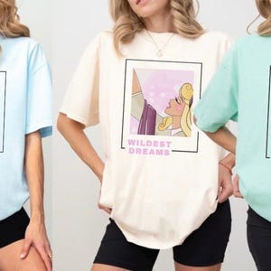 May include: Three light-colored t-shirts with different Disney princess graphics and text. The first shirt is light blue with a graphic of Cinderella and the text "Meet me at Midnight". The second shirt is cream with a graphic of Aurora from Sleeping Beauty and the text "Wildest Dreams". The third shirt is light green with a graphic of Ariel from The Little Mermaid and the text "But Daddy I Love Him".
