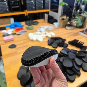 Könnte beinhalten: Ein schwarz-weißes Objekt mit Wabenmuster wird in der Hand gehalten. Das Objekt ist von anderen ähnlichen schwarzen Objekten auf einem Holztisch umgeben. Weitere Objekte sind im Hintergrund sichtbar.