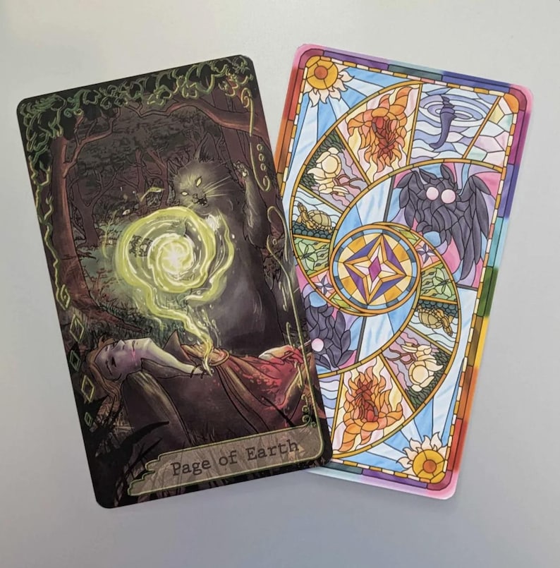 May include: A tarot card with a dark green, brown, and gold illustration of a cat and a person with a glowing orb. The card is labeled "Page of Earth". The back of the card is a colorful stained glass design.
