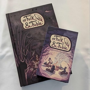 May include: A purple and black hardcover book titled "Of Folk & Fable" with a smaller purple and black card game box with the same title. The card game box has the text "A collaborative tarot project" on the front.