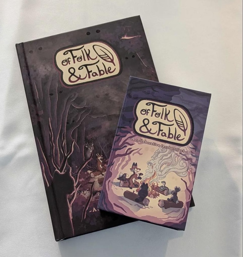 May include: A purple and black hardcover book titled "Of Folk & Fable" with a smaller purple and black card game box with the same title. The card game box has the text "A collaborative tarot project" on the front.