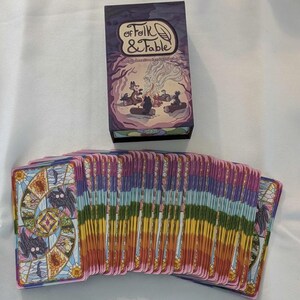 May include: A deck of Folk & Fable oracle cards with a purple box. The box has a colorful illustration of a fox, a wolf, and a bear sitting around a campfire. The cards have a stained glass design with a rainbow color scheme.