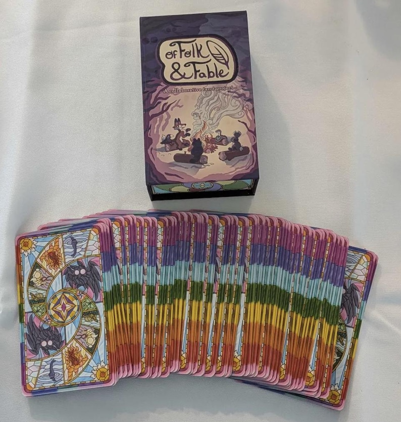 May include: A deck of Folk & Fable oracle cards with a purple box. The box has a colorful illustration of a fox, a wolf, and a bear sitting around a campfire. The cards have a stained glass design with a rainbow color scheme.