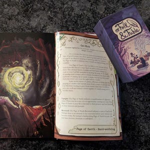 May include: A purple card deck box with the text "Folk & Fable" and a feather illustration. The box is open and shows a card with a purple background and a cat-like creature with a yellow aura. The card text reads "Cat-Sith" and "Page of Earth - Hard-working".