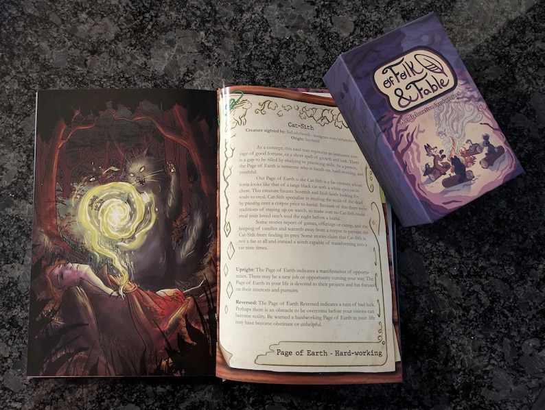 May include: A purple card deck box with the text "Folk & Fable" and a feather illustration. The box is open and shows a card with a purple background and a cat-like creature with a yellow aura. The card text reads "Cat-Sith" and "Page of Earth - Hard-working".