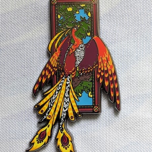 May include: A colorful enamel pin featuring a stylized phoenix with red, orange, yellow, and black feathers. The phoenix is perched on a branch in a stained glass window with a blue sky and green leaves.
