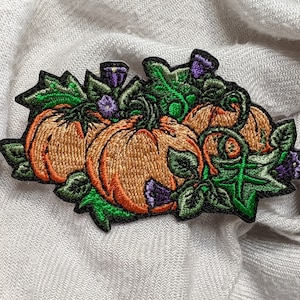 Może przedstawiać: Naszywka haftowana przedstawiająca trzy pomarańczowe dynie z zielonymi winoroślami i fioletowymi kwiatami. Dynie są ułożone w grupie i mają realistyczny wygląd.