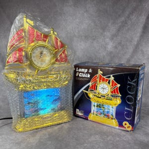 May include: A gold-colored ship-shaped lamp and clock combination featuring red sails and a circular clock face. The base contains a decorative underwater scene. The item is wrapped in clear bubble wrap next to its original cardboard packaging box.