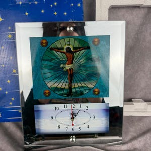 May include: A rectangular wall clock with a mirrored frame featuring a central circular image of the crucifixion of Jesus against a teal sunburst background. Below the image is an oval clock face with black numerals and hands on a white background.