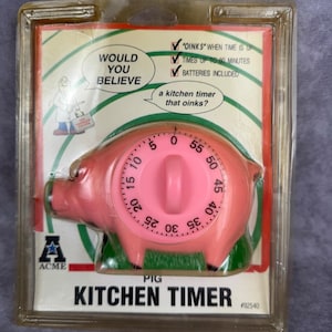 May include: A pink pig-shaped kitchen timer in its original plastic packaging. The timer features a circular dial with numbers from 0 to 55. The packaging includes the text "WOULD YOU BELIEVE a kitchen timer that oinks?" and "KITCHEN TIMER".