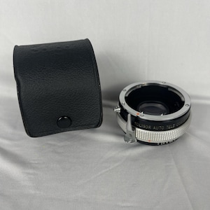 Puede incluir: Un estuche negro de cuero para lentes de cámara y una lente Poligor Auto Tele Converter plateada y negra. La lente tiene un anillo de ajuste plateado con números y el texto "POLIGOR AUTO TELE CONV". El estuche es rectangular con cierre de botón.