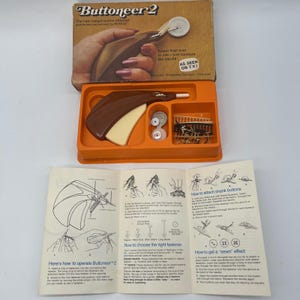 Op de afbeelding: Vintage Buttoneer 2 knopenaanzetkit in de originele doos. De kit bevat de bruine en crèmekleurige tool, knopen en instructies. Op de doos staat "The new instant button attacher" en "AS SEEN ON T.V."
