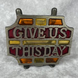 Pode incluir: Placa decorativa de metal com as palavras "GIVE US THIS DAY" em letras vermelhas. A placa apresenta vitrais em tons de âmbar, amarelo e vermelho. Possui uma moldura de metal prateado e pés pequenos.