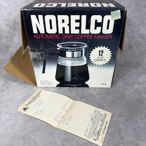May include: A vintage Norelco automatic drip coffee maker in its original box. The box is black with white text and features an image of the 12-cup carafe. A receipt is in front of the box.