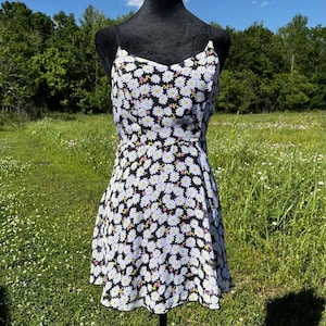 Puede incluir: Vestido con estampado floral en blanco y negro con escote en V y tirantes finos. El vestido presenta un estampado de margaritas con pétalos blancos, centros amarillos y detalles rosas. El vestido se exhibe en un maniquí negro.