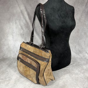 May include: A brown patchwork shoulder bag featuring multiple exterior zippered compartments with dark brown trim and two long shoulder straps. The bag is constructed from various shades of tan and brown suede panels, showcasing a textured surface.