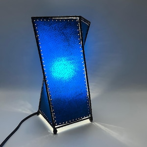 May include: A blue, geometric table lamp with a black frame. The lamp has a twisted design with a bright blue light emanating from within. The lamp is connected to a black power cord.