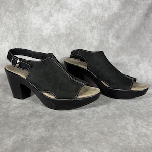 May include: A pair of black suede open-toe sandals featuring a thick platform sole, a sturdy block heel, and an adjustable ankle strap with a silver-toned metal buckle. The interior footbed is a light beige color with visible white contrast stitching.
