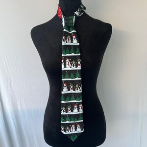 May include: A black necktie featuring a repeating pattern of snowmen, green Christmas trees, and snowflakes. The snowmen wear hats and scarves in various colors. The tie is displayed on a black mannequin.