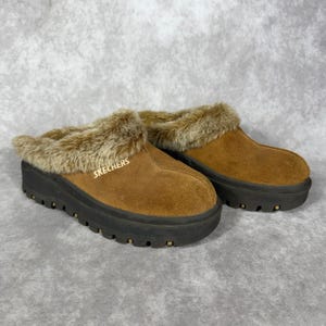 Könnte beinhalten: Ein Paar braune Wildleder-Clogs zum Hineinschlüpfen mit einer dicken, dunkelbraunen Profilsohle und einem flauschigen, hellbraunen Kunstfellbesatz am Kragen. Das Wort SKECHERS ist in kleinen, hellen Buchstaben an der Seite des Schuhs eingestickt.