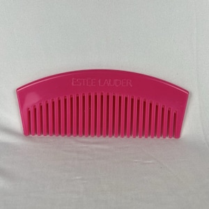 May include: A bright pink comb with the text "ESTÉE LAUDER" printed on the top. The comb has a curved top and many thin teeth. The comb is made of plastic and is set against a white background.