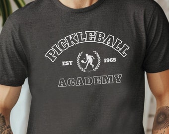 Może przedstawiać: Szary t-shirt z białym nadrukiem graficznym, na którym widnieje napis "Pickleball Academy" z wieńcem laurowym i tekstem "Est 1965".