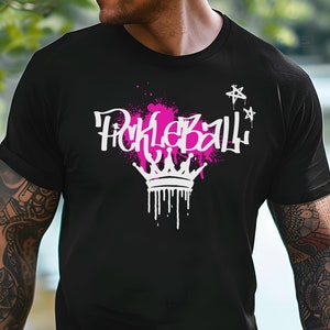 May include: Black t-shirt with a white and pink graphic that says "Pickleball" with a crown design. The text has a graffiti-style font with a pink paint splatter effect. Two white stars are above the text.