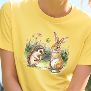 Puede incluir: Una camiseta amarilla con una ilustración de dibujos animados de un erizo y un conejo jugando al pickleball. El erizo sostiene una pala de pickleball y el conejo sostiene una pala de pickleball. Hay una pelota de pickleball en el aire entre ellos.