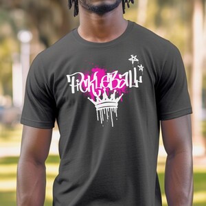 May include: Dark gray t-shirt featuring the word "Pickleball" in white graffiti-style lettering with a pink splatter design. A white crown with dripping paint is below the word, and white stars are above.