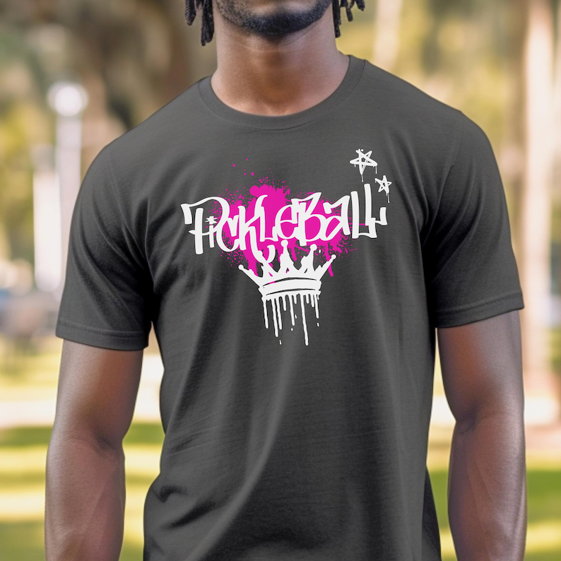 May include: Dark gray t-shirt featuring the word "Pickleball" in white graffiti-style lettering with a pink splatter design. A white crown with dripping paint is below the word, and white stars are above.