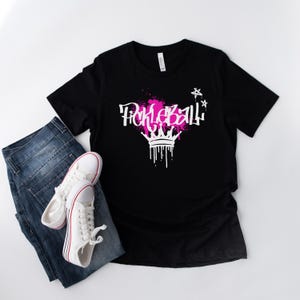 May include: Black t-shirt with the word "Pickleball" in white graffiti-style font, accented with pink splatters and a white crown design. The shirt is paired with blue jeans and white sneakers with red stripes.