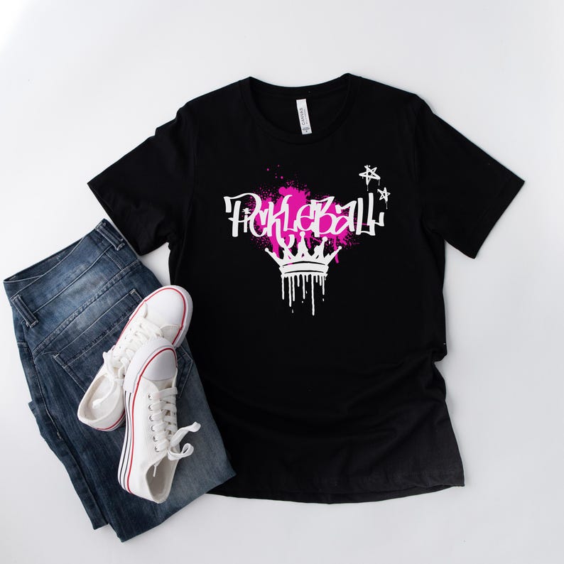May include: Black t-shirt with the word "Pickleball" in white graffiti-style font, accented with pink splatters and a white crown design. The shirt is paired with blue jeans and white sneakers with red stripes.