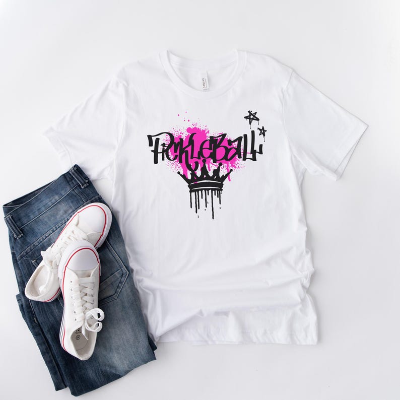 May include: White t-shirt with a black and pink graphic that says "Pickleball" with a crown design. The shirt is paired with blue jeans and white sneakers.